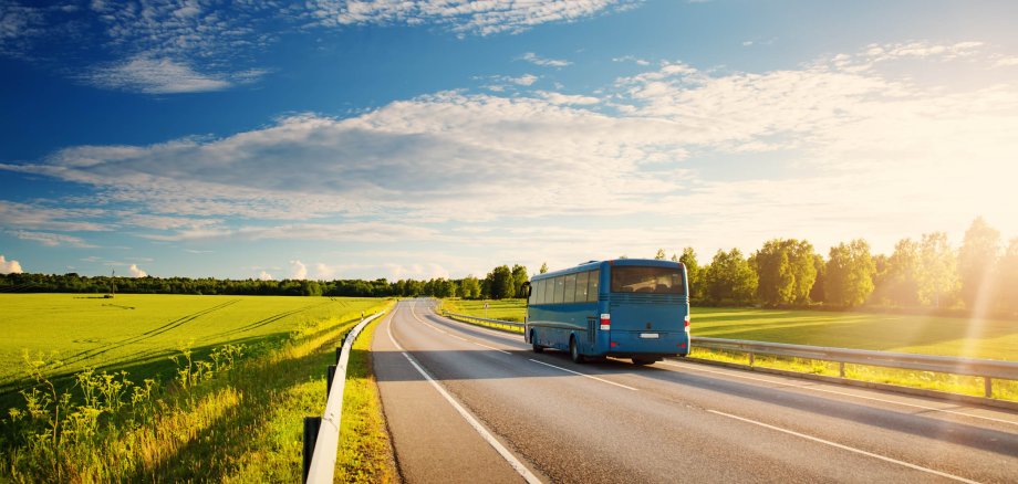 Ein blauer Bus fährt eine Landstraße entlang. Ringsum sind bewachsene Felder und ein blauer Himmel mit leichten Wolken zu sehen