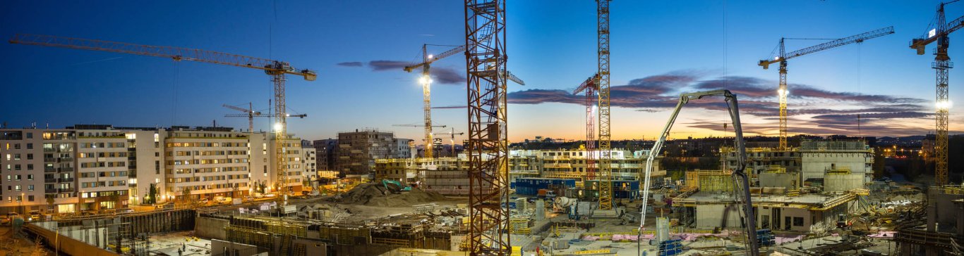 Zahlreiche Baukräne, teilweise beleuchtet, vor abendlichem Himmel in einer Großbaustelle. Im Hintergrund sind fertiggestellte Hochhäuser zu sehen.